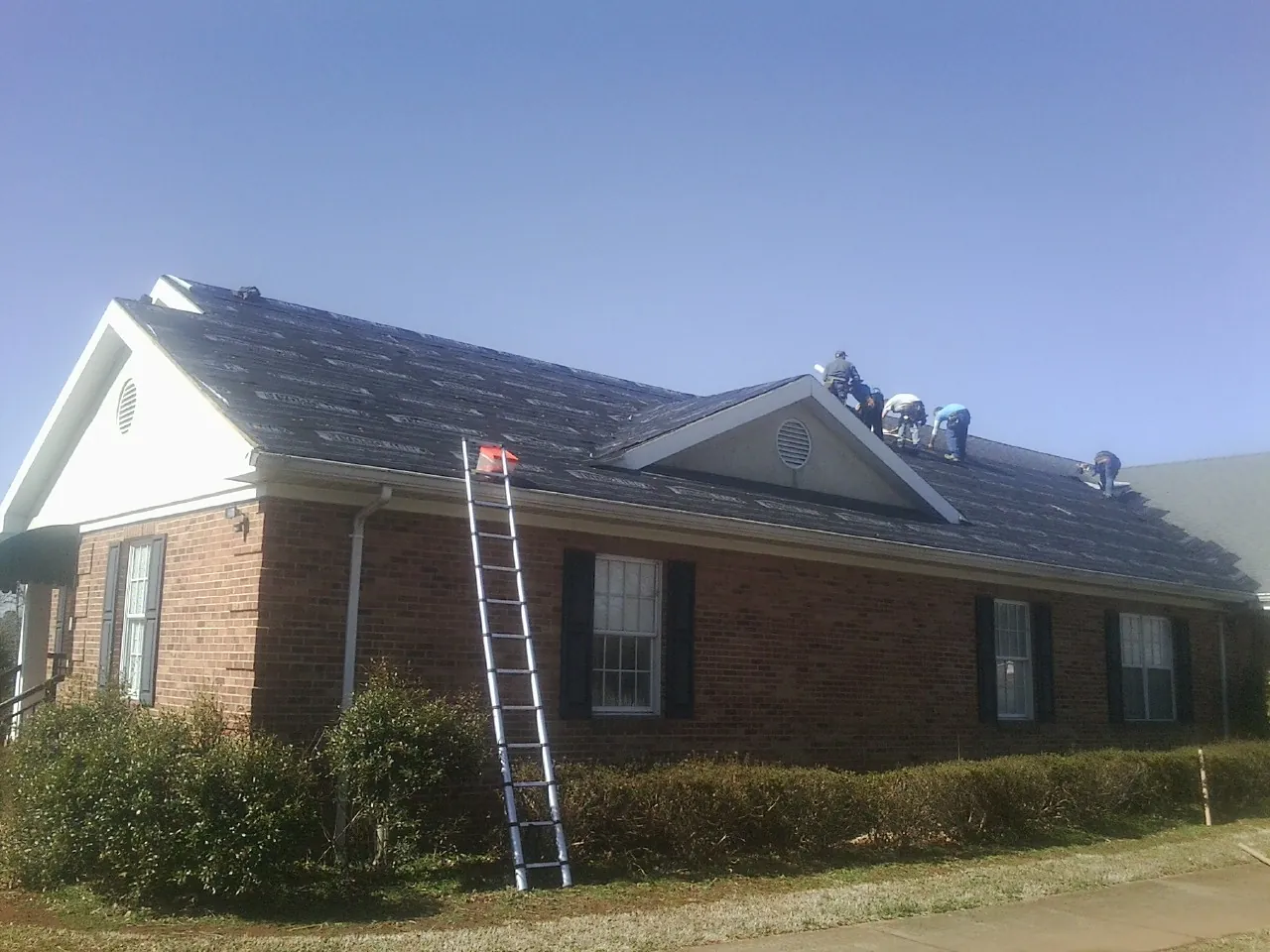 Crew tearing shingles off a brick ranch roof with a ladder set — Metal Roof Repair work by our crew in Bethany